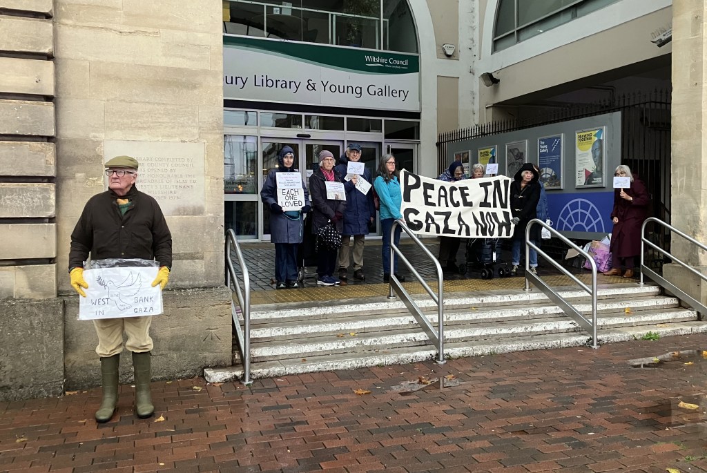 A group of protesters holding signs advocating for peace in Gaza, gathered outside a public library and gallery. One individual, dressed in rubber boots and gloves, stands to the side holding a sign that reads 'WEST BANK IN GAZA'.