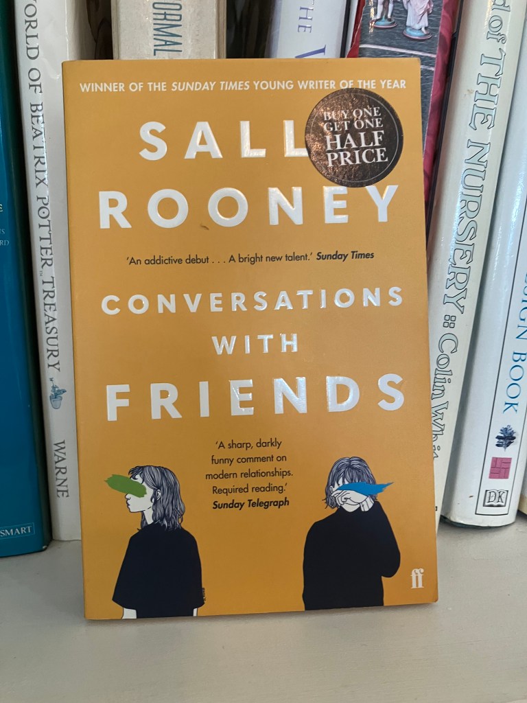 A close-up of the book 'Conversations with Friends' by Sally Rooney, featuring an orange cover with white text and illustrations of two figures with obscured faces. A sticker indicating a buy one get one half price offer is also present.