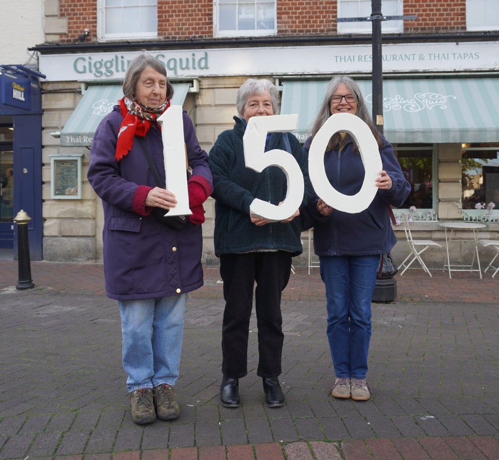 Three women standing outdoors, holding large numbers '1', '5', and '0' to signify '150'. They are in front of a restaurant with outdoor seating.