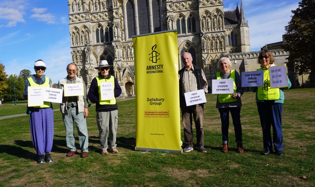 Group of protesters holding signs advocating for Uyghur rights in front of a historic building, with an Amnesty International banner in the background.